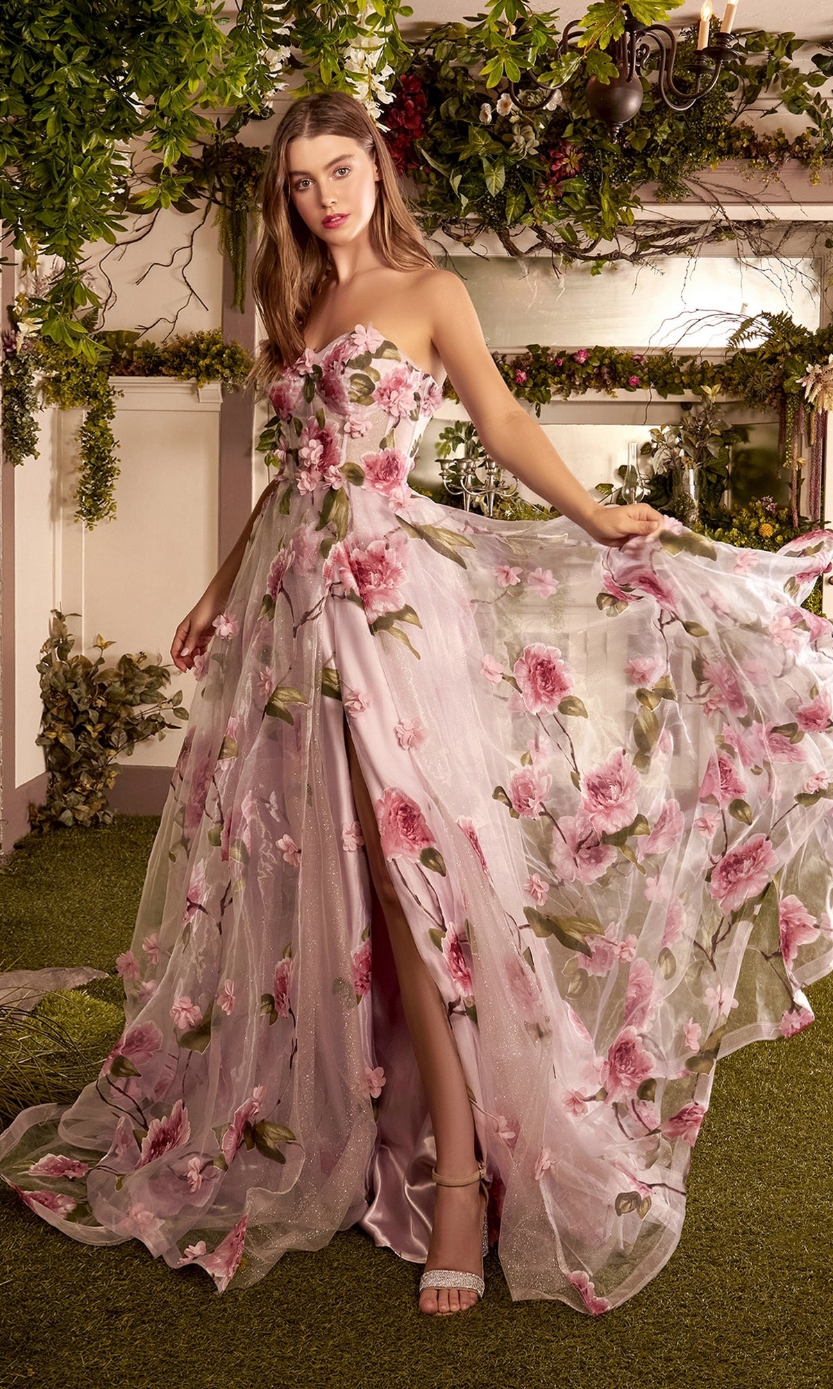 A woman stands on grass, holding out the skirt of her Floral-Print Strapless Long Prom Ball Gown A1035, which features pink and green floral patterns and a corset-style waist.